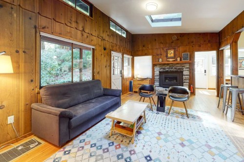 Cozy living room with wooden walls, a fireplace, a sofa, and a colorful rug, featuring large windows and natural light.
