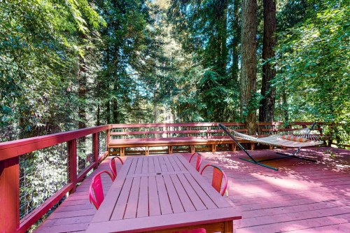 A wooden deck surrounded by tall trees, featuring a dining table and a hammock for relaxation.