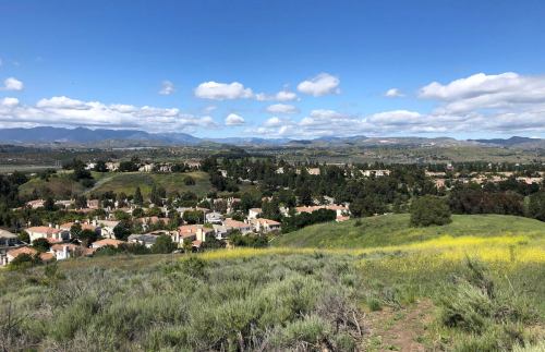 A scenic view of a suburban area with houses, green hills, and mountains under a partly cloudy sky.