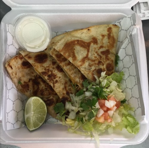 A takeout container with a quesadilla, salad, lime wedge, and a small cup of sauce.