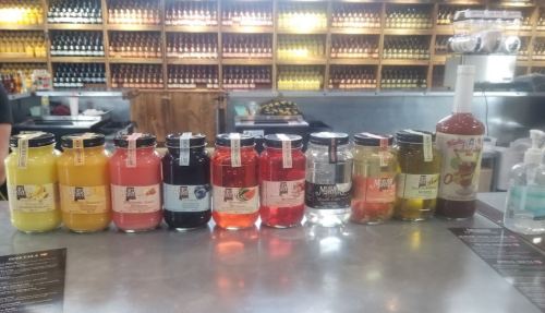 A variety of colorful bottled drinks lined up on a counter, with labels and a backdrop of shelves filled with more bottles.