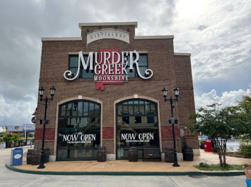 Exterior of the Murder Creek Moonshine distillery, featuring large windows and a "NOW OPEN" sign.