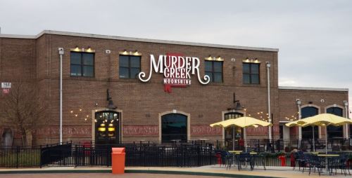Exterior of a building with a sign that reads "Murder Creek Moonshine," featuring outdoor seating and a cloudy sky.