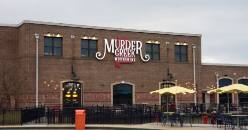 Exterior of a brick building with a sign that reads "Murder Creek Moonshine," featuring outdoor seating and umbrellas.