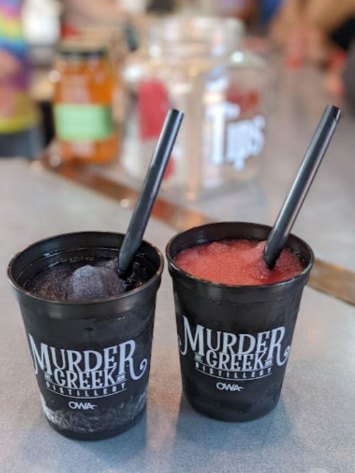 Two cups from Murder Creek Distillery filled with colorful frozen drinks and black straws on a bar counter.