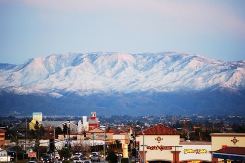 Snow-capped mountains rise above a suburban landscape at dusk, with shops and buildings in the foreground.