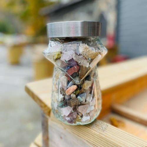 A glass jar filled with colorful stones and crystals, sitting on a wooden surface outdoors.