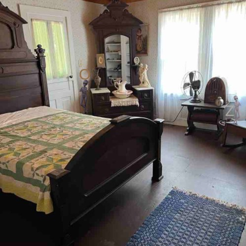 A vintage bedroom featuring a wooden bed, dresser, and decorative items, with sunlight streaming through the windows.