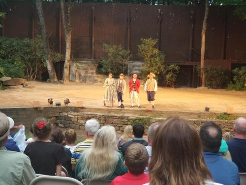 A theater performance with three actors on stage, viewed by an audience seated in front. Trees and a rustic backdrop are visible.