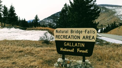 Sign for Natural Bridge Falls Recreation Area in Gallatin National Forest, with mountains and snow in the background.