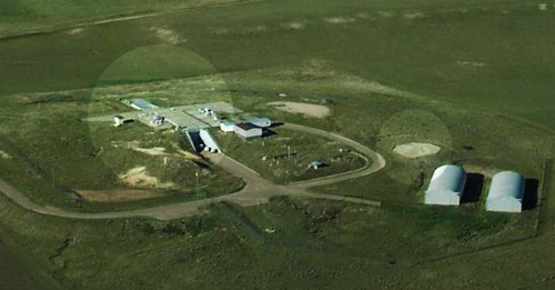 Aerial view of a remote facility surrounded by open fields, featuring several buildings and storage structures.