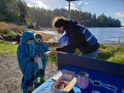 A person in a blue vest assists two children in blue jackets by a lakeside, with trees and a cloudy sky in the background.