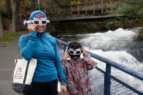 A woman and a child wear fun sunglasses by a river, smiling and posing for the camera.