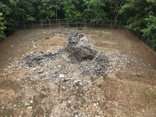 A cleared, rocky area surrounded by a fence, with a large mound of dirt and stones in the center, set in a wooded environment.