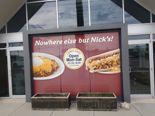 Sign on a storefront featuring "Nowhere else but Nick's!" with images of food and summer hours listed.