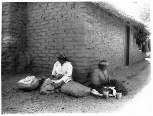 Two women sit on stones outside a mud-brick house, one weaving and the other preparing food.