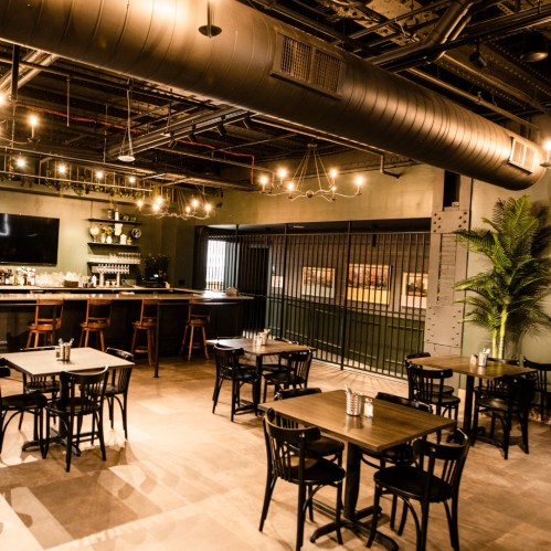 A modern restaurant interior featuring wooden tables, chairs, a bar area, and industrial-style decor.
