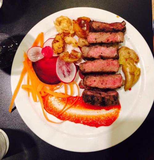 A plate of grilled steak slices with colorful vegetables, garnished with sauce and crispy onion rings.