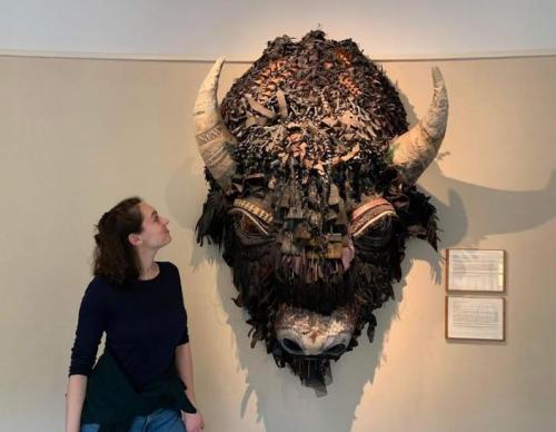 A person gazes up at a large, textured bison head sculpture made of various materials on a wall.