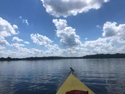 A kayak's bow on calm water, with a bright blue sky filled with fluffy clouds in the background.