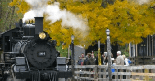 A vintage steam train emits smoke as it approaches a wooden platform surrounded by autumn trees and onlookers.