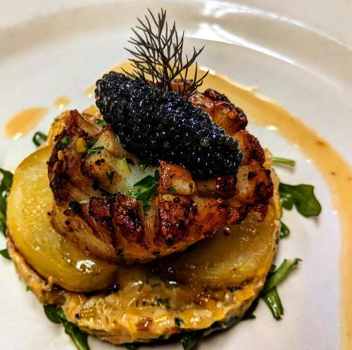 A gourmet dish featuring grilled seafood atop sliced potatoes, garnished with caviar and herbs on a creamy base.