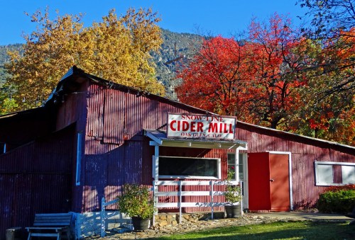 A rustic cider mill with a red exterior, surrounded by colorful autumn foliage and mountains in the background.