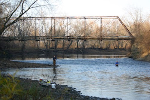 Two people fishing in a river with a rusty, old bridge in the background and bare trees lining the shore.