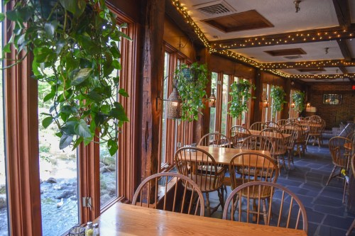 Cozy restaurant interior with wooden tables, chairs, and greenery, featuring large windows overlooking a stream.