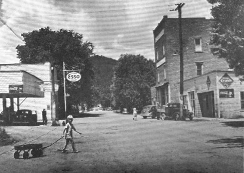 A black-and-white photo of a small town street with vintage cars, a child pulling a wagon, and old storefronts.