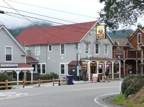 A quaint gray building with a red roof, featuring a sign and outdoor seating, set in a small town.