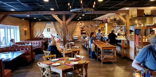 Cozy restaurant interior with wooden decor, tables set for dining, and staff preparing food in an open kitchen.