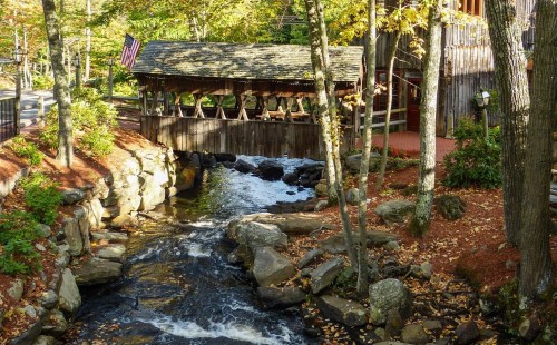 A scenic view of a covered bridge over a stream, surrounded by autumn foliage and rocky banks.