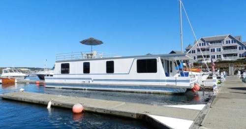 A white houseboat docked at a marina on a sunny day, with other boats and a building in the background.