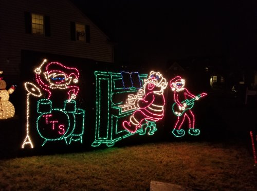 Colorful Christmas lights depict a band with a drummer, keyboardist, and guitarist in a festive outdoor display.