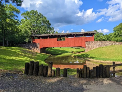 A red covered bridge spans a calm pond, surrounded by green grass and trees under a partly cloudy sky.