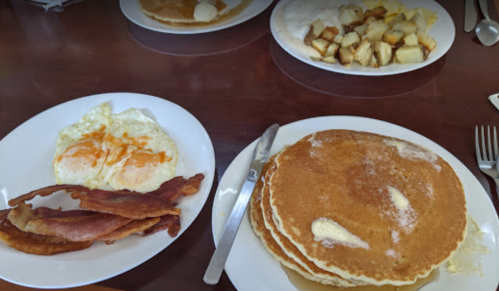 A plate of fluffy pancakes with butter and syrup, alongside crispy bacon and eggs, with a side of home fries.