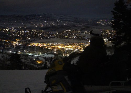A person on a snowmobile overlooks a snowy landscape and a lit-up town at night.