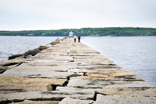 Two people walk along a rocky pier extending into calm waters, with a lighthouse visible in the distance.