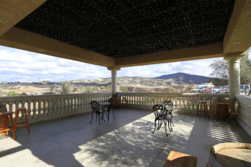 A covered patio with chairs and tables, overlooking rolling hills under a cloudy sky. String lights adorn the ceiling.