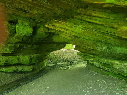 A moss-covered rock formation with a natural arch, leading to a lush green landscape beyond.