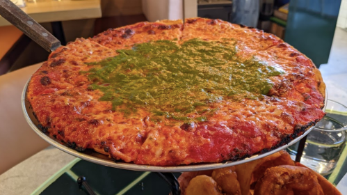 A large pizza topped with melted cheese and a vibrant green sauce, served on a metal tray.