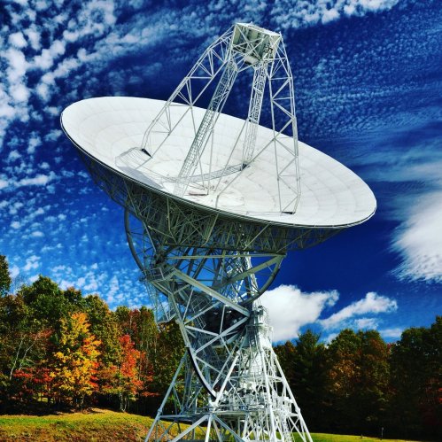 A large satellite dish stands against a blue sky with clouds, surrounded by colorful autumn trees.