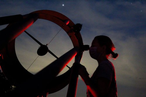 A silhouette of a person wearing a mask, looking through a telescope against a twilight sky.