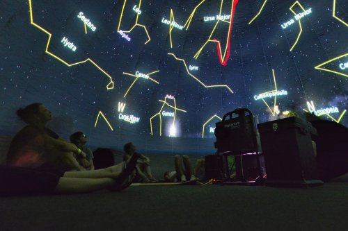 A group of people lying on the floor inside a planetarium, gazing at a starry sky with constellations projected above.