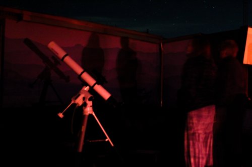 A telescope set up outdoors at night, with shadows of people observing the stars in the background.