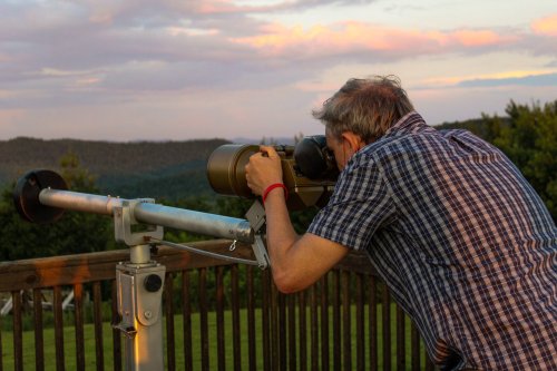 A person in a plaid shirt uses a telescope to observe a scenic landscape at sunset.