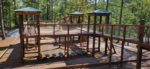 A wooden playground with climbing structures and nets, surrounded by trees and gravel paths.