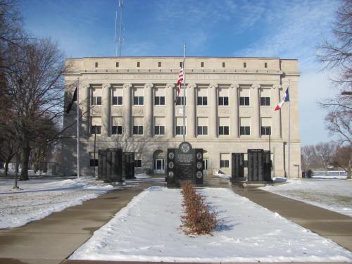 A stone courthouse with flags in front, surrounded by snow and trees, under a clear blue sky.