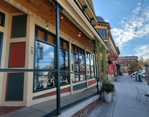 A charming bakery with large windows, inviting signage, and a sunny street view lined with shops and greenery.